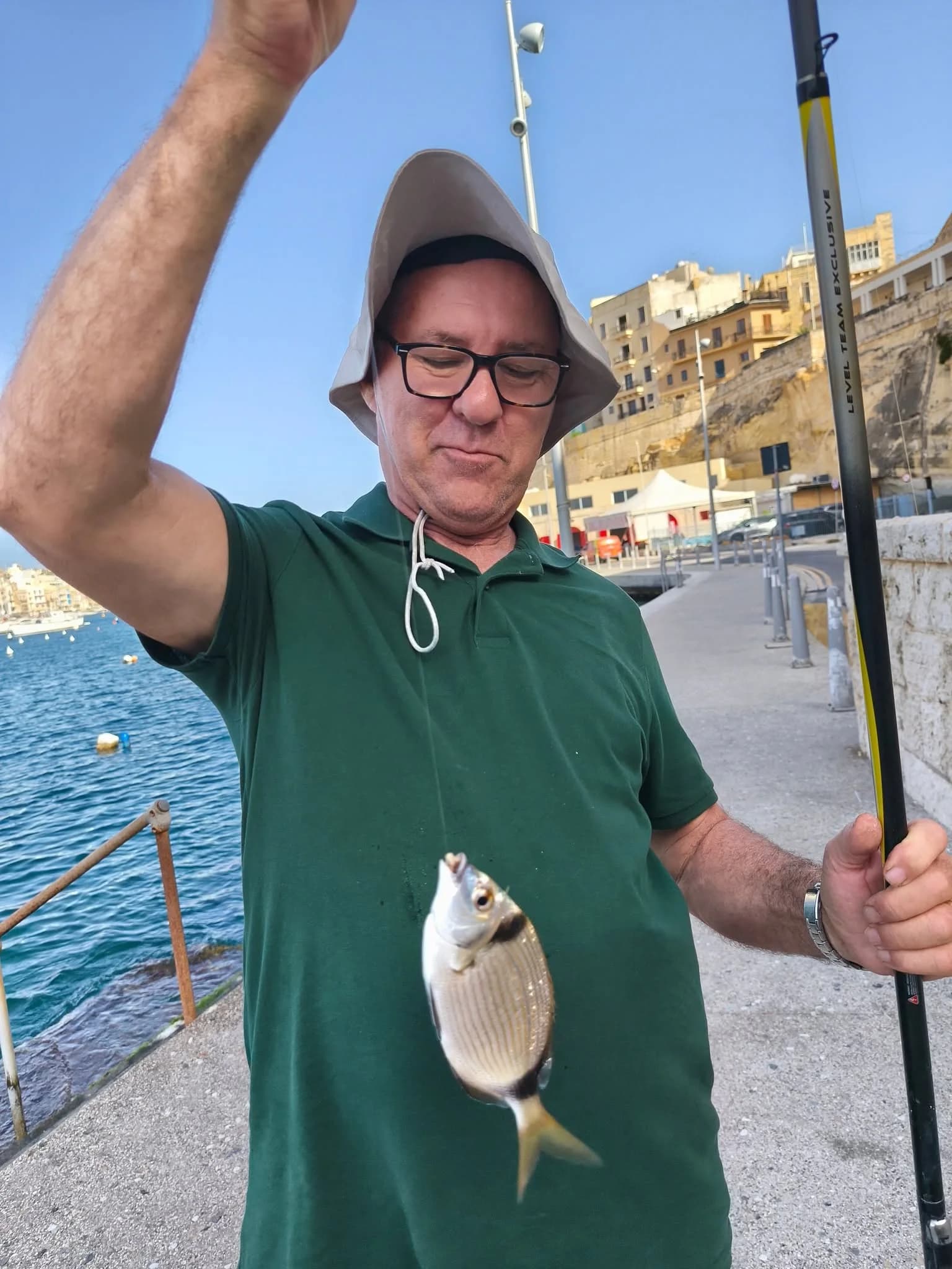 An angler with glasses holding a fishing pole and catch beside the water