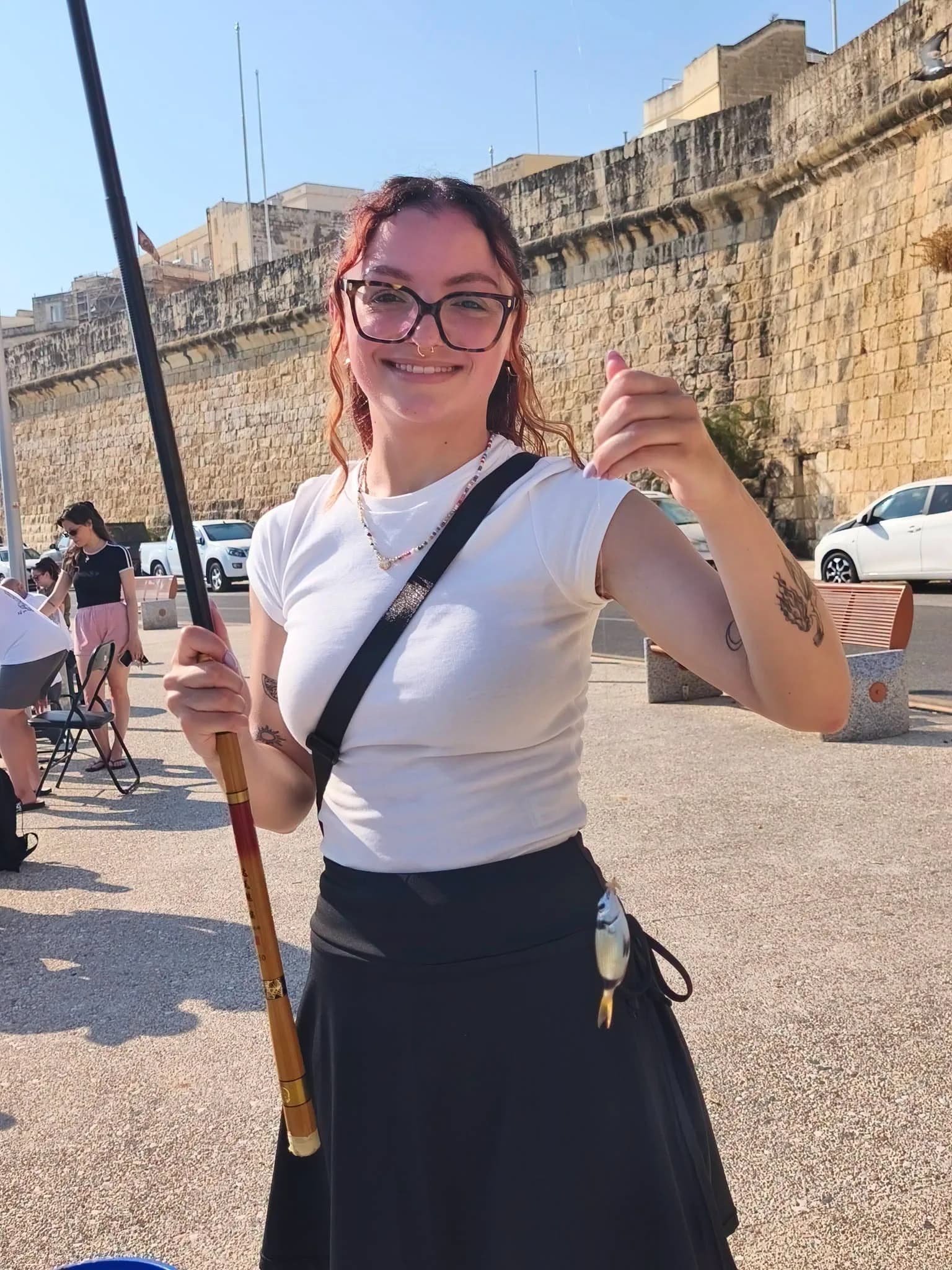 An angler with glasses holding a pole and fish in front of the waterfront buildings