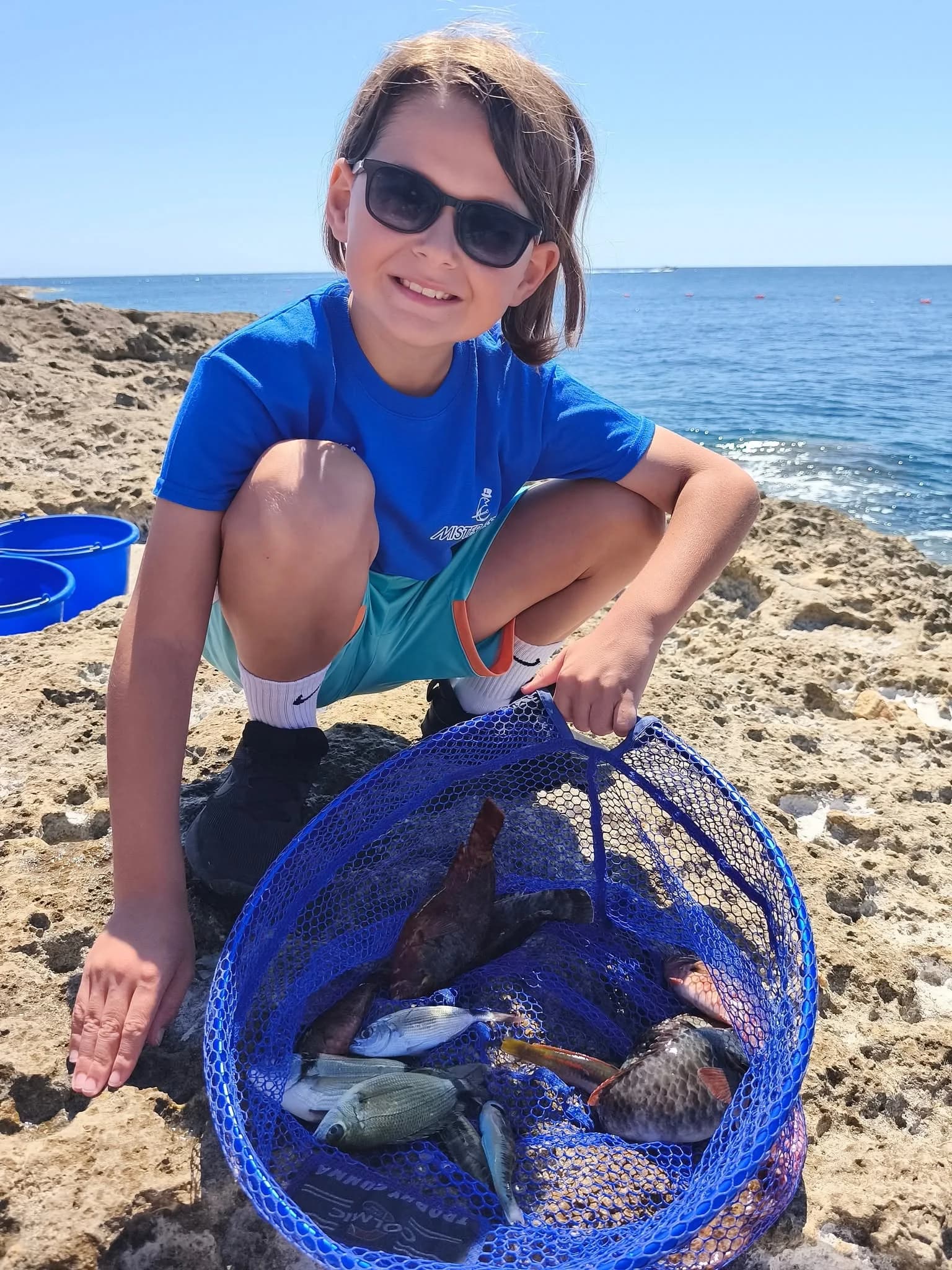 A smiling young angler kneeling beside a net and fishing bucket