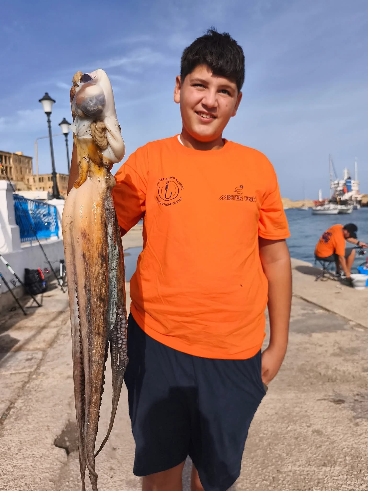 A young angler in an orange shirt holding a fish at the waterfront
