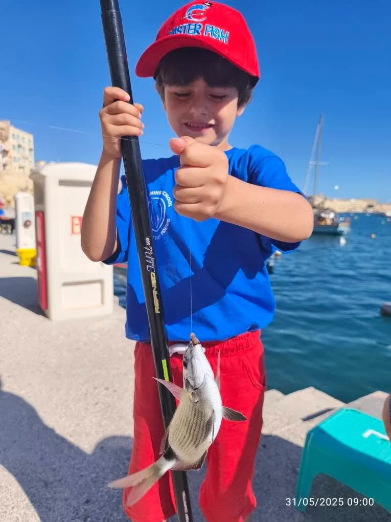 A young angler in a blue shirt and red cap holding a fish at the harbour