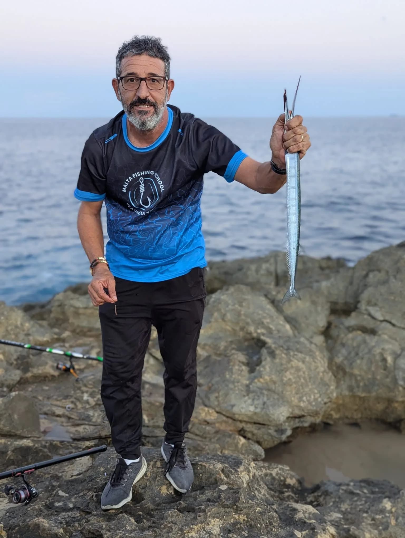 An angler in a Malta Fishing School shirt holding a catch beside the sea
